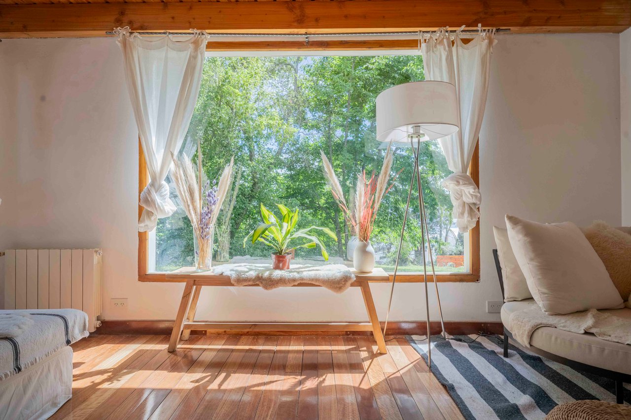 Bedroom with a large tree-framed window, sheer curtains, and warm timber flooring.