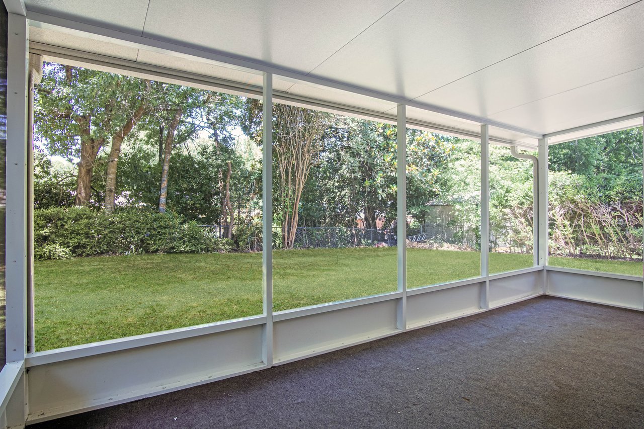 Enclosed porch with large windows overlooking a lush, green backyard. Trees and bushes surround the space, providing a serene, natural view.