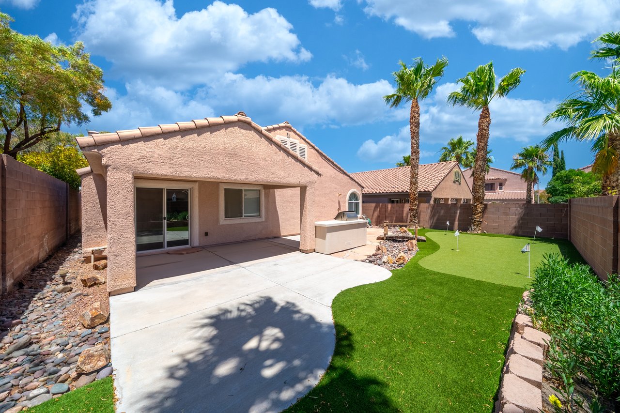 Home’s backyard with a covered patio, artificial turf putting green, palm trees, and outdoor counter.