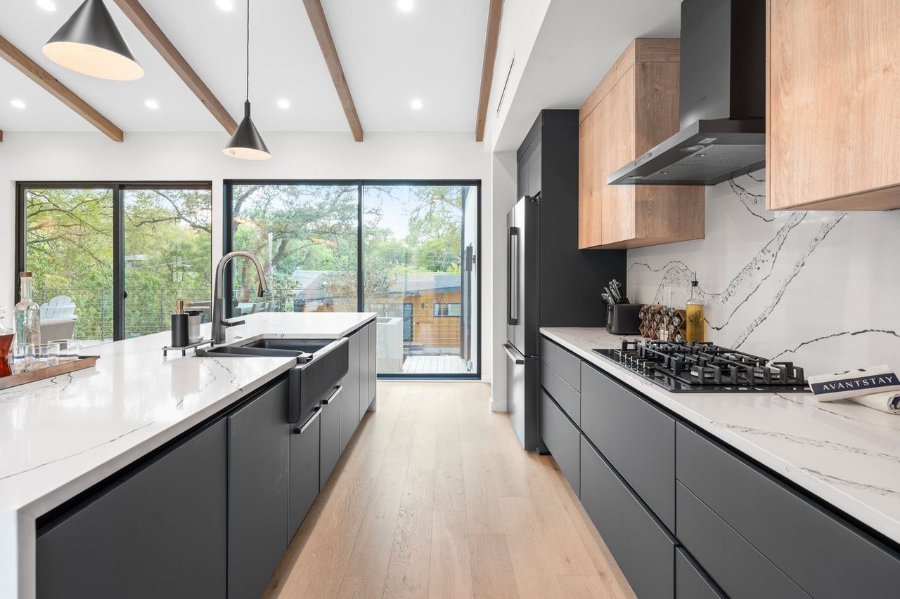 Modern kitchen with a large double-sink island, dark cabinets, wood beams, and glass doors overlooking trees.