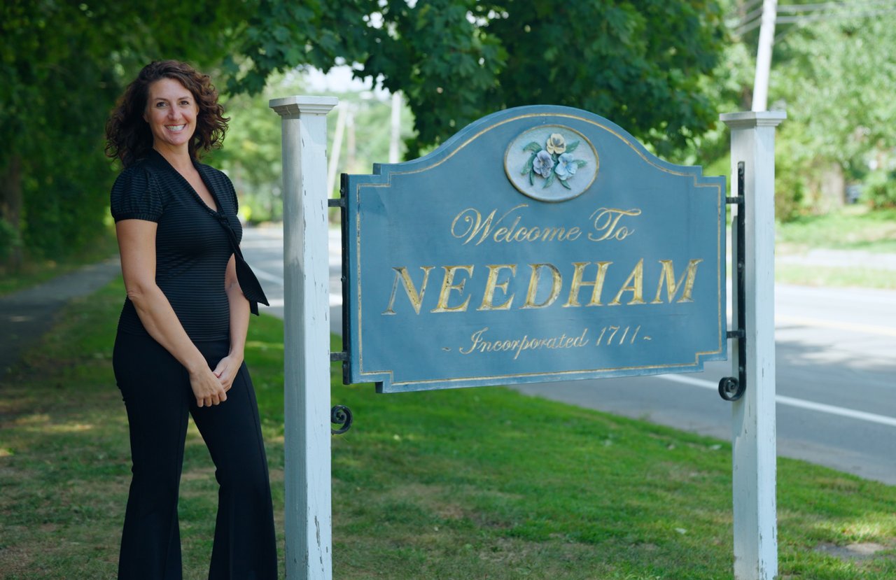 A woman stands beside a blue Needham town sign beneath leafy trees.