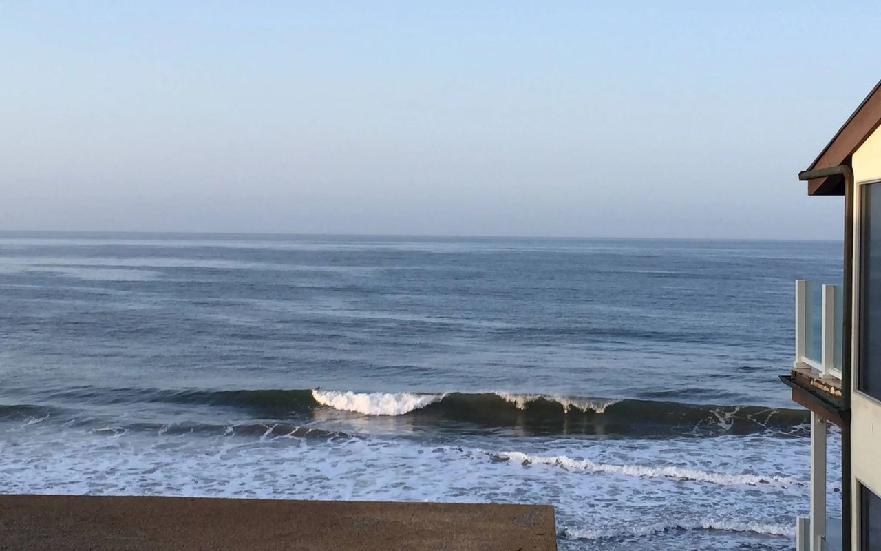 Ocean waves break below a coastal home, with part of its balcony visible at the right.