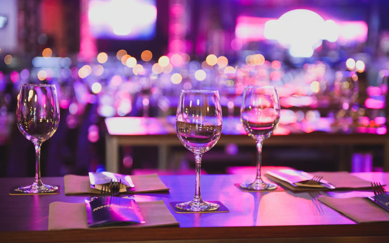 A restaurant table set with wine glasses and cutlery, featuring a moody atmosphere with vibrant pink and purple lights.