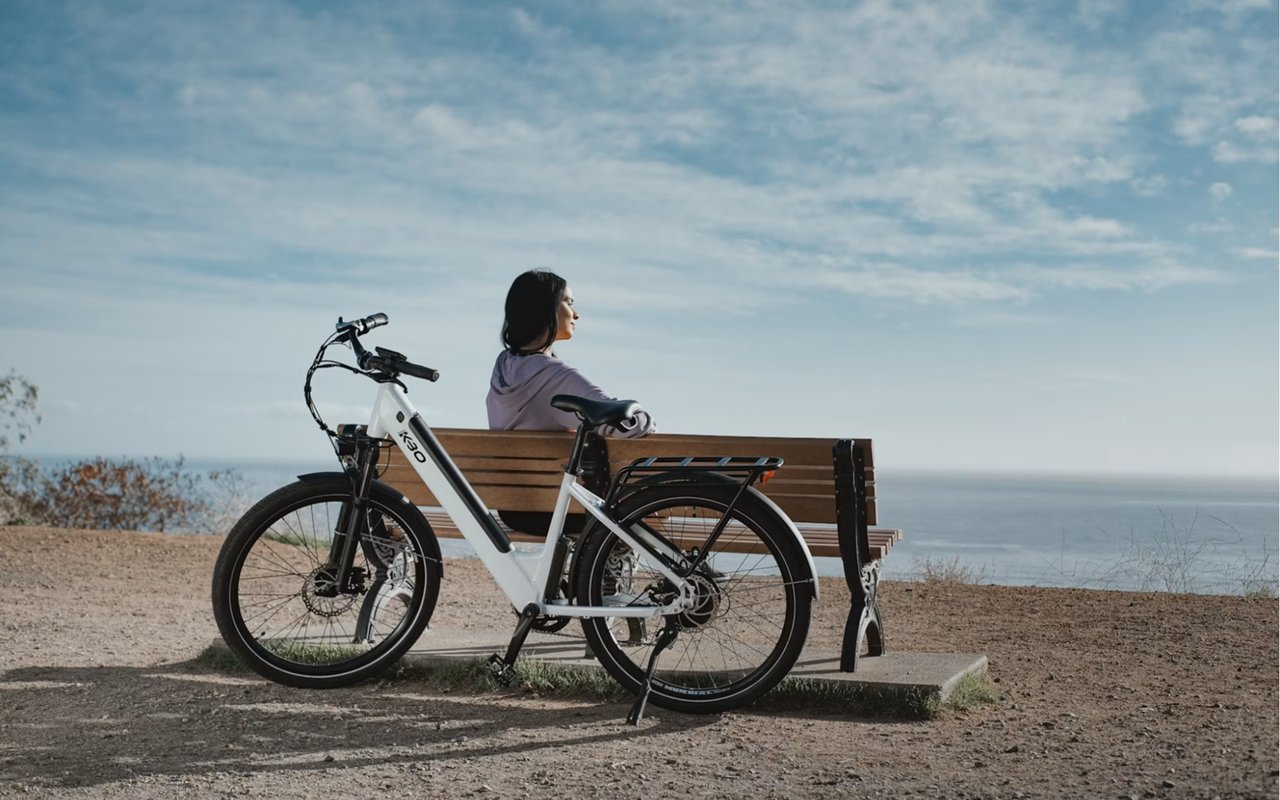 Woman sitting on a lakeside bench beside a white electric bicycle, looking out over the water.