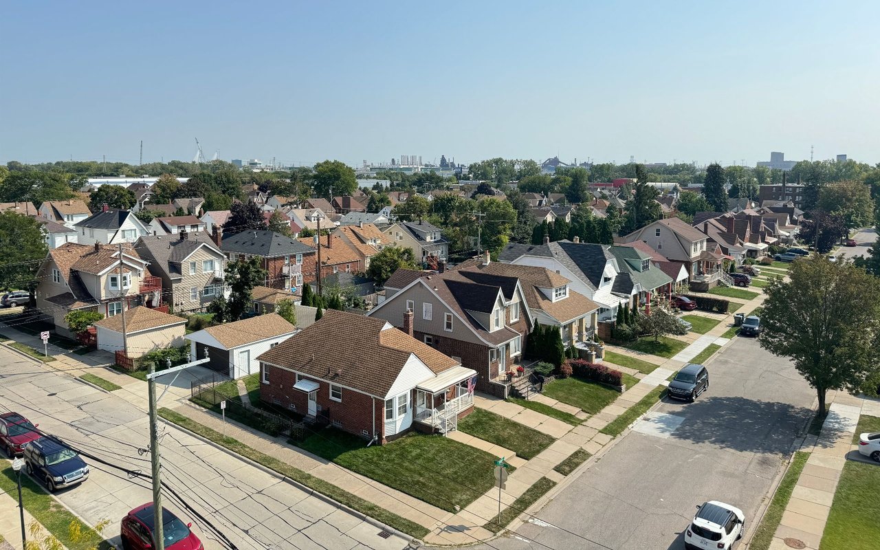 Aerial view of a residential neighborhood with tree-lined streets and an industrial skyline in the distance.