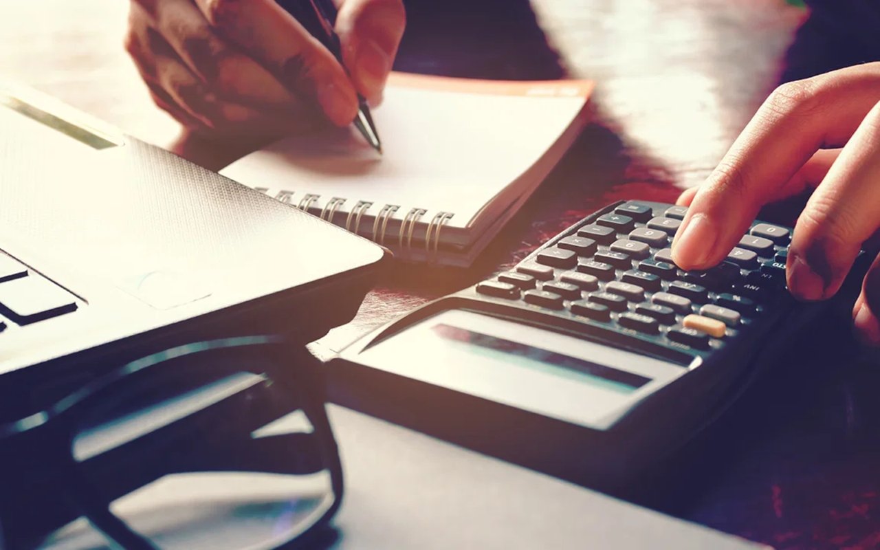 Hands write in a notebook and use a calculator beside paperwork and eyeglasses.