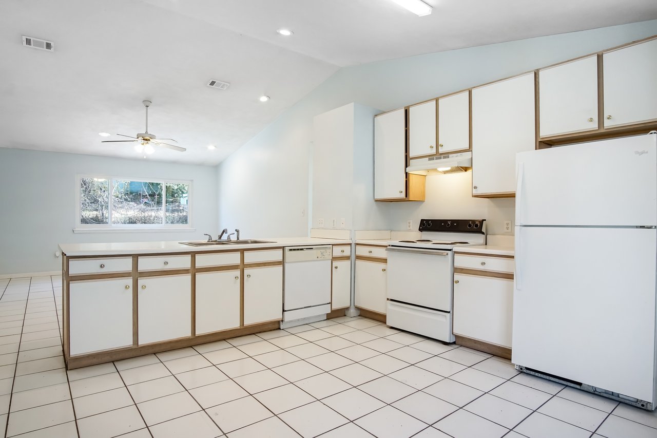 Spacious kitchen with white cabinets, tile flooring, and a white fridge. A ceiling fan above and large window create a bright, airy atmosphere.