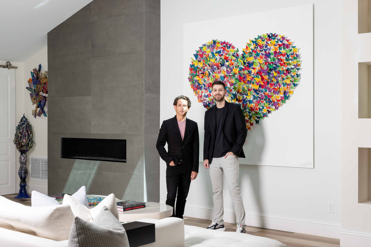 Two men pose in a modern living room beneath colorful heart-shaped artwork.