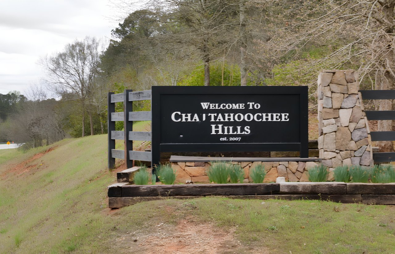 Chattahoochee Hills entrance sign beside a stone pillar and wooded roadside.