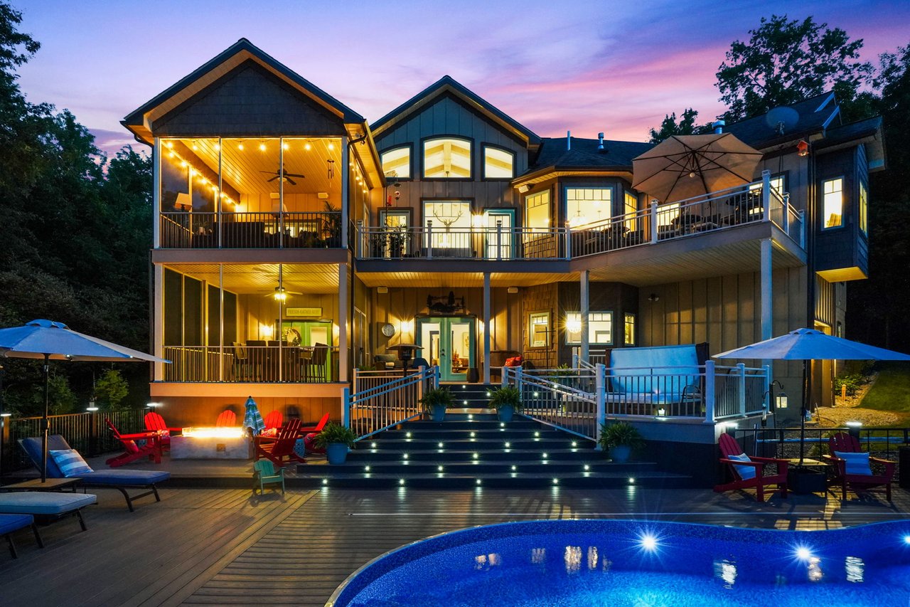 Twilight view of a custom luxury lakefront estate at 23018 Loop Road in Senecaville, Ohio, featuring illuminated multi-level decks, screened porches, resort-style pool, outdoor fire pit, and panoramic views overlooking Seneca Lake.