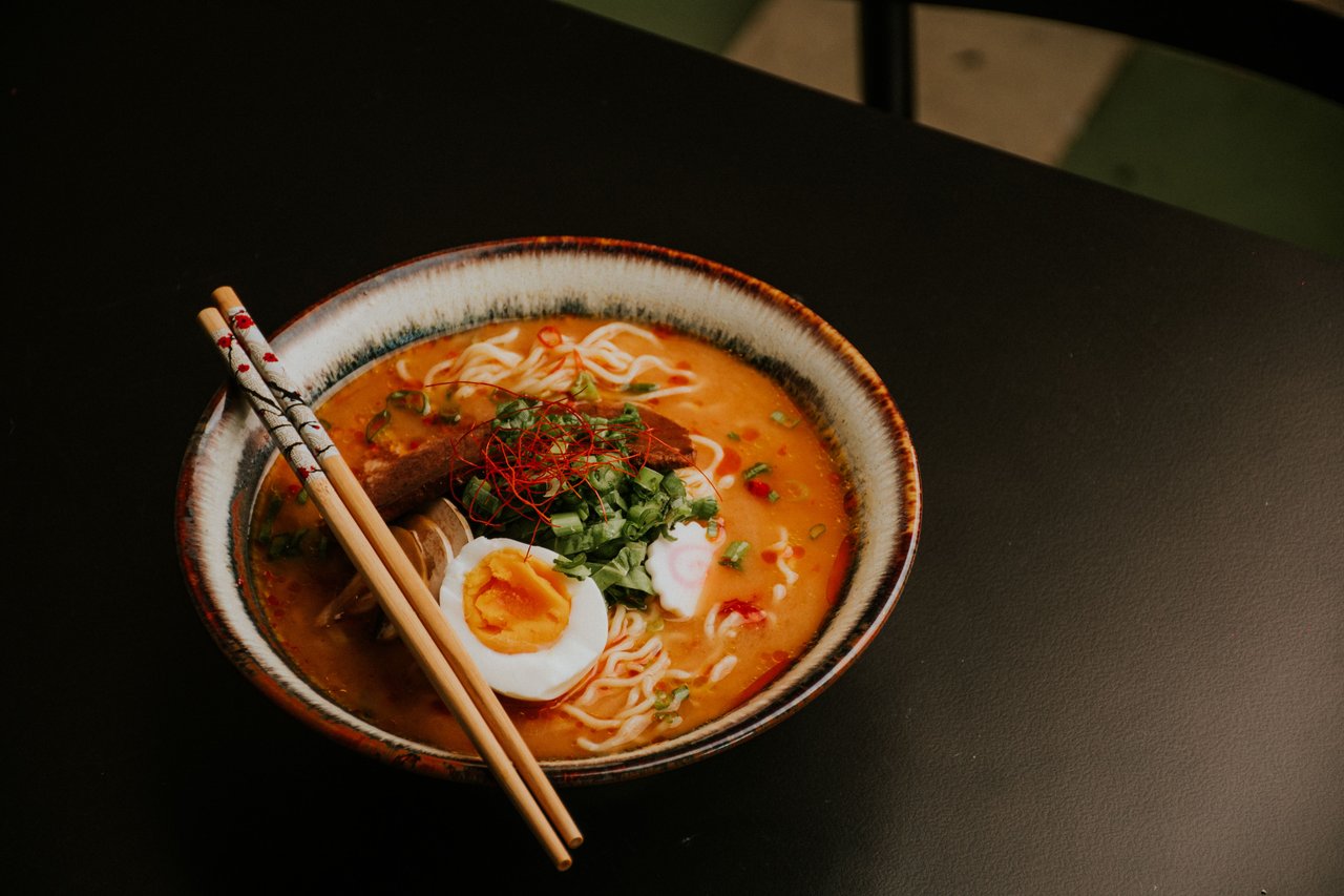Ramen with orange broth, soft-boiled egg, greens, chili threads, and chopsticks.