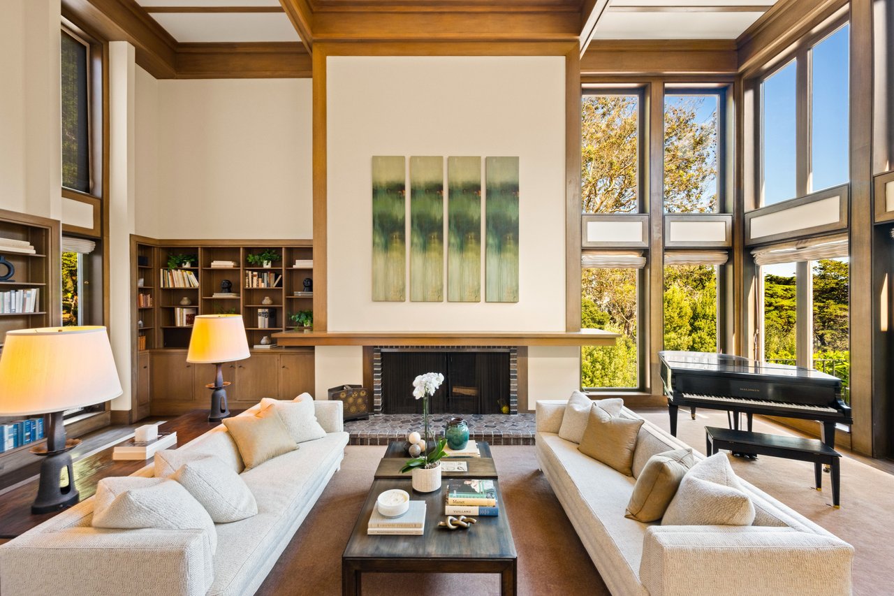 Living room with a tall stone fireplace, vaulted wood ceiling, and expansive windows beside a grand piano.