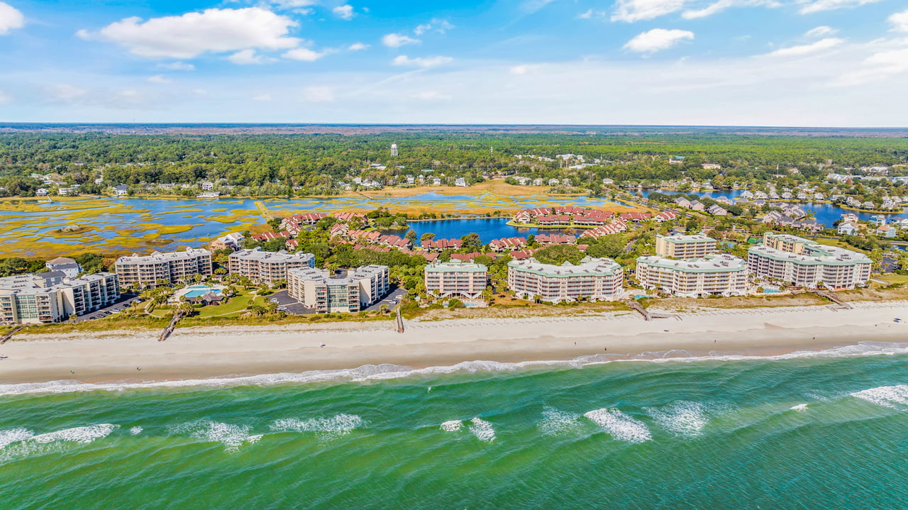 Beachfront condominiums line a sandy shore, with ocean waves and marshland behind them.
