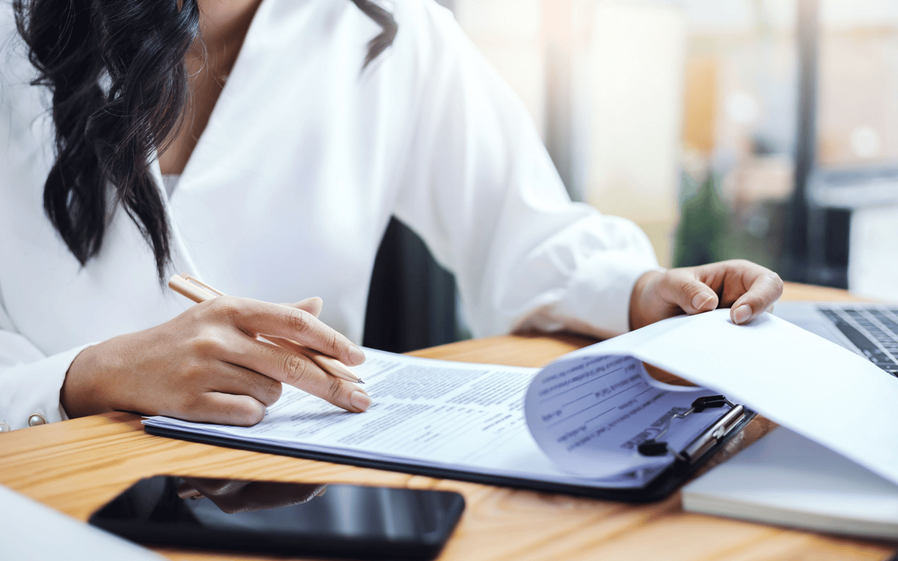 Woman reviewing paperwork with a pen beside a laptop and smartphone