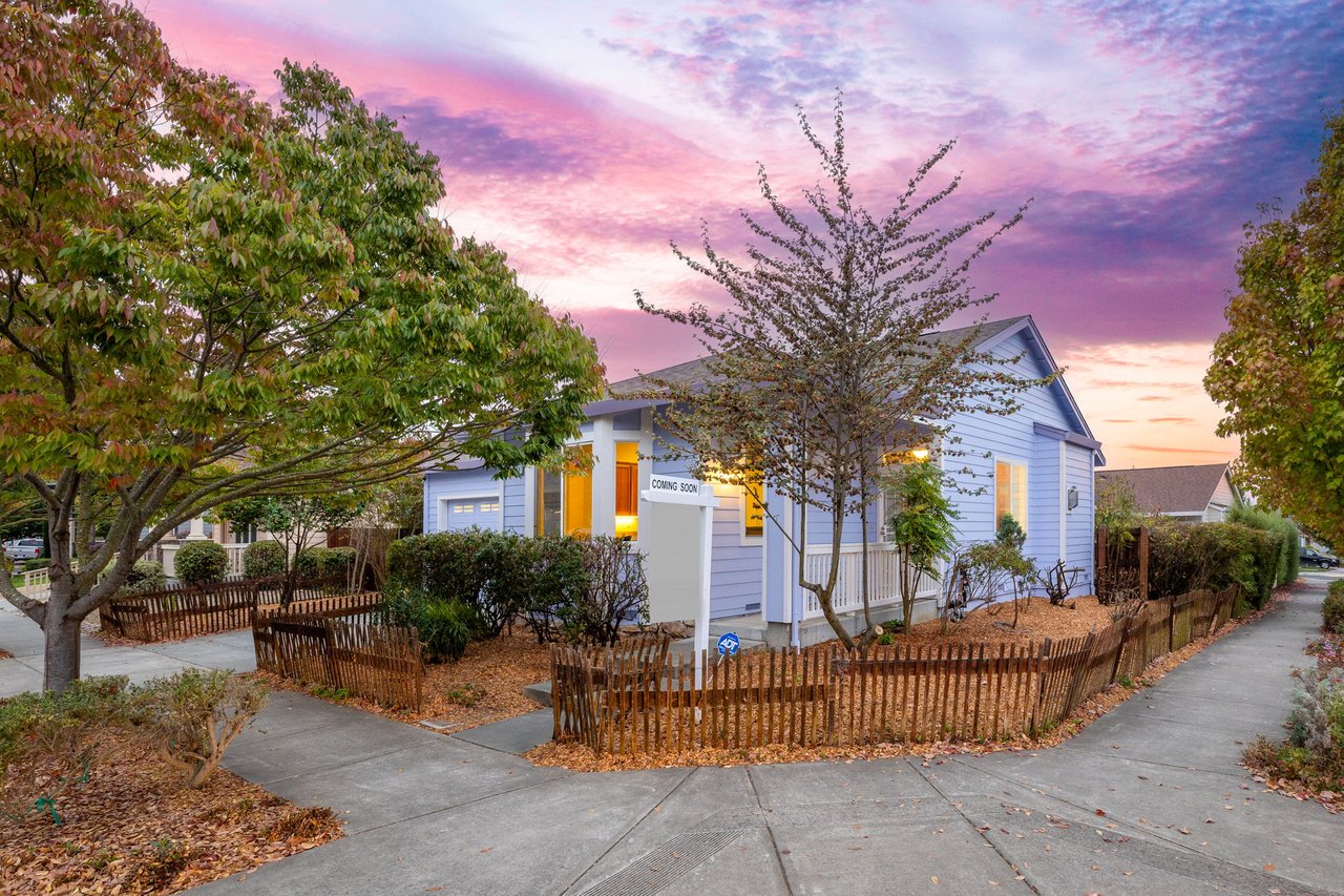 Blue-sided house with a white porch and fenced yard beneath a vivid pink sunset sky.