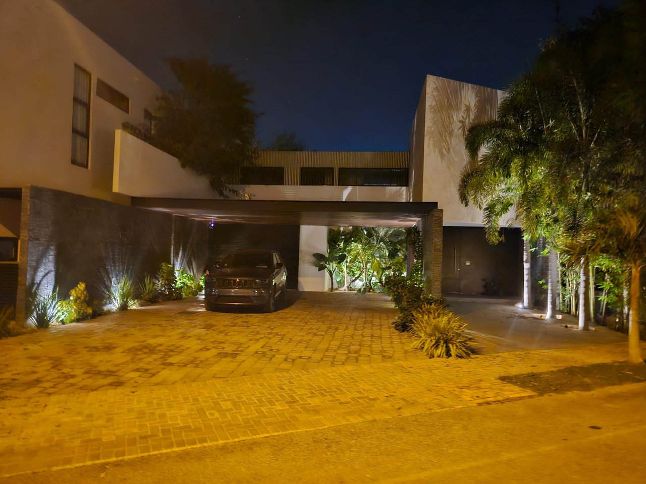 Modern residence at night with a covered driveway, tropical landscaping, and a warmly lit paved entrance.