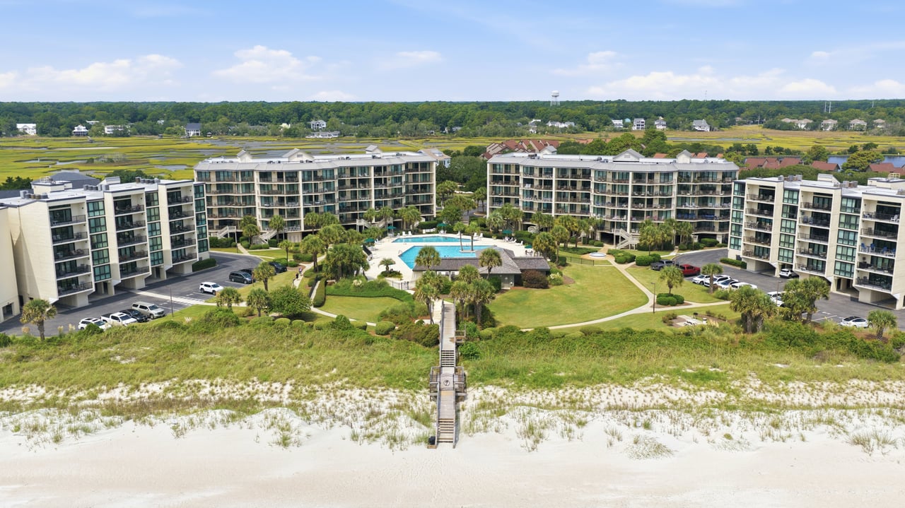 Beachfront condominium complex surrounding a pool, with a boardwalk crossing dunes to the sandy beach.