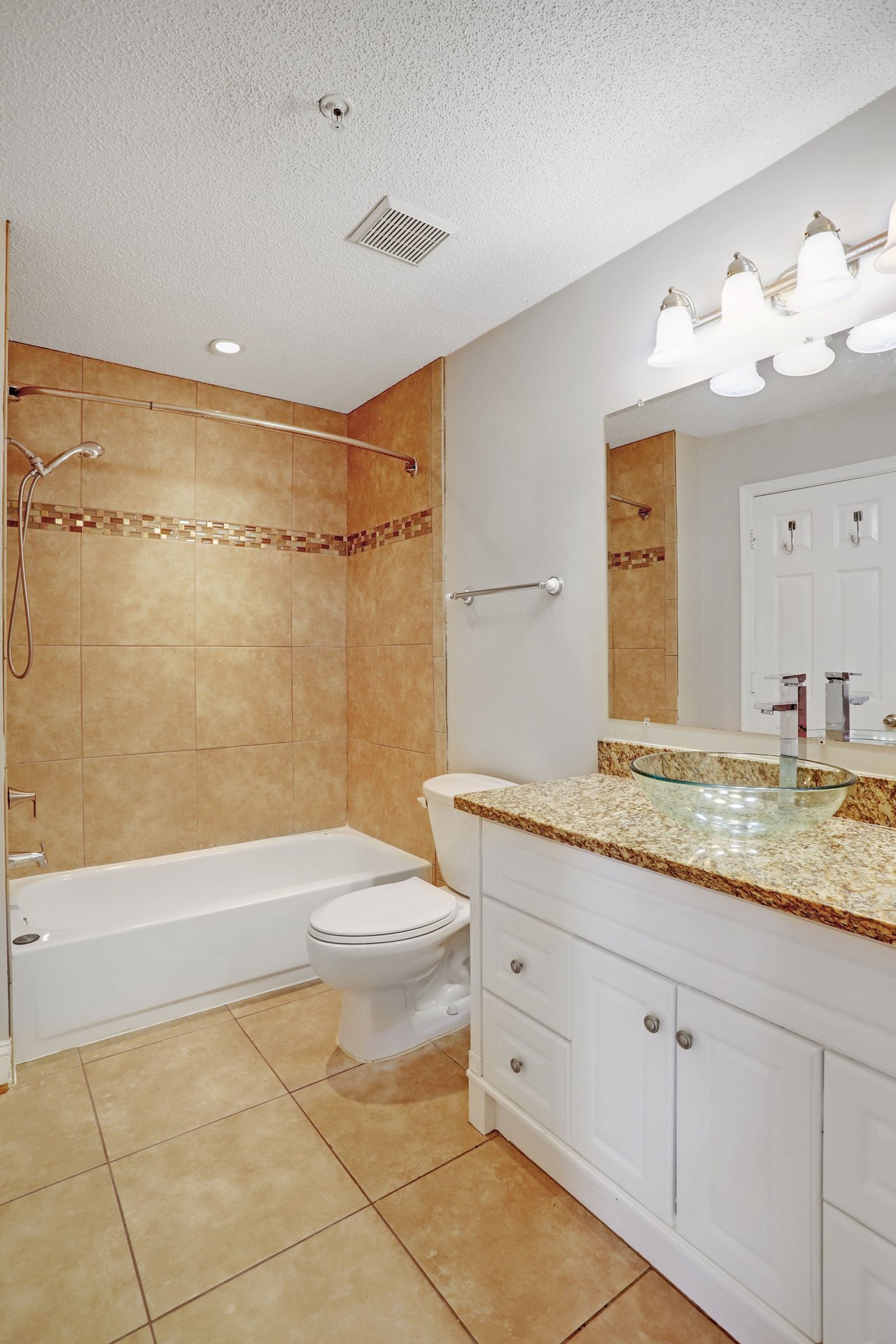 Bathroom with tan tiled tub and shower, white vanity, granite counter, and glass vessel sink.