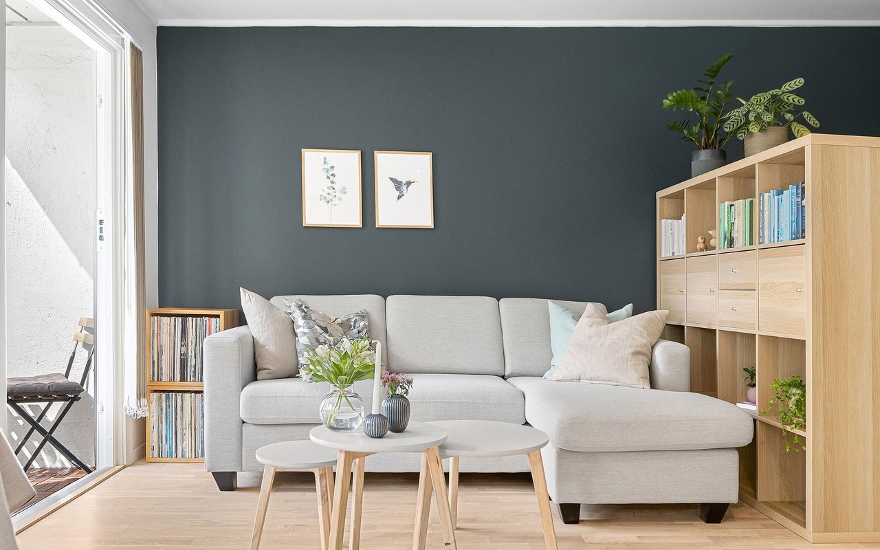 Living room with a gray sectional, dark teal accent wall, pale wood shelving, and bright balcony doors.