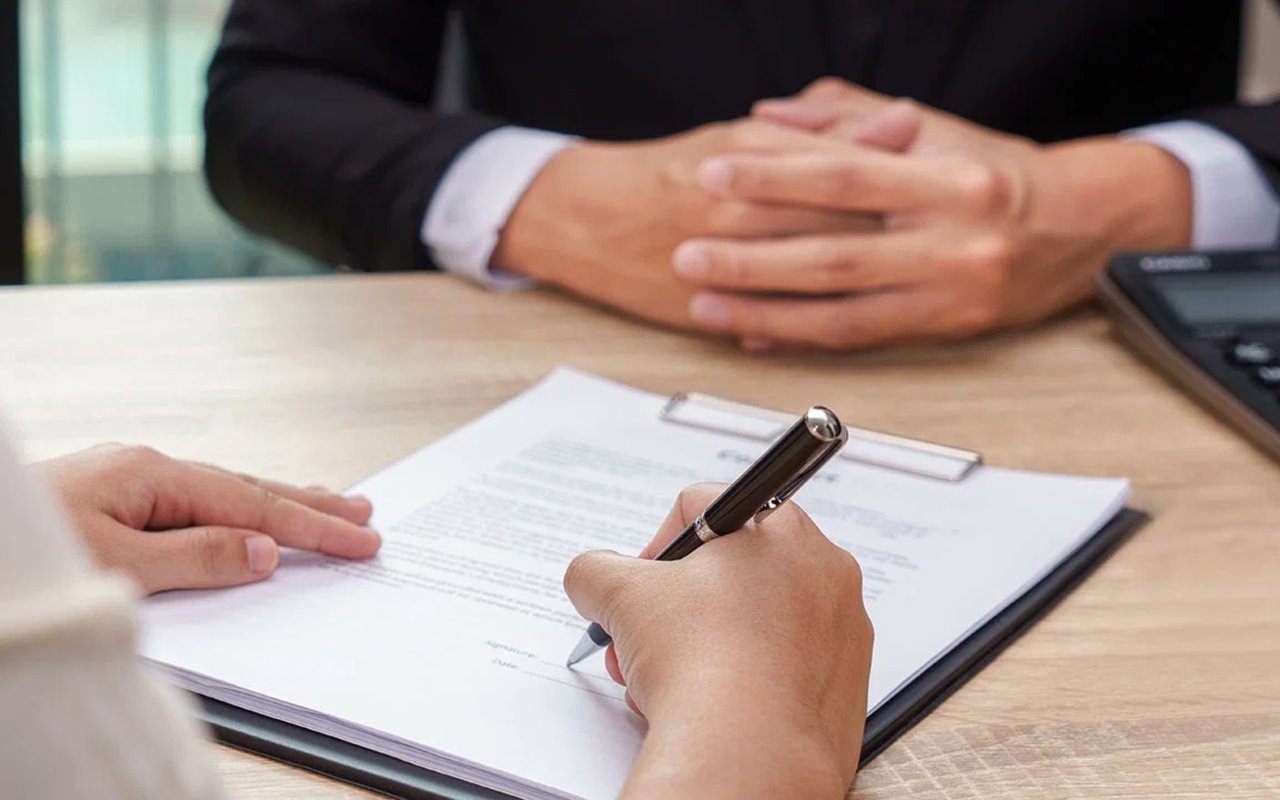 A person signs mortgage paperwork while a suited person sits across the desk.