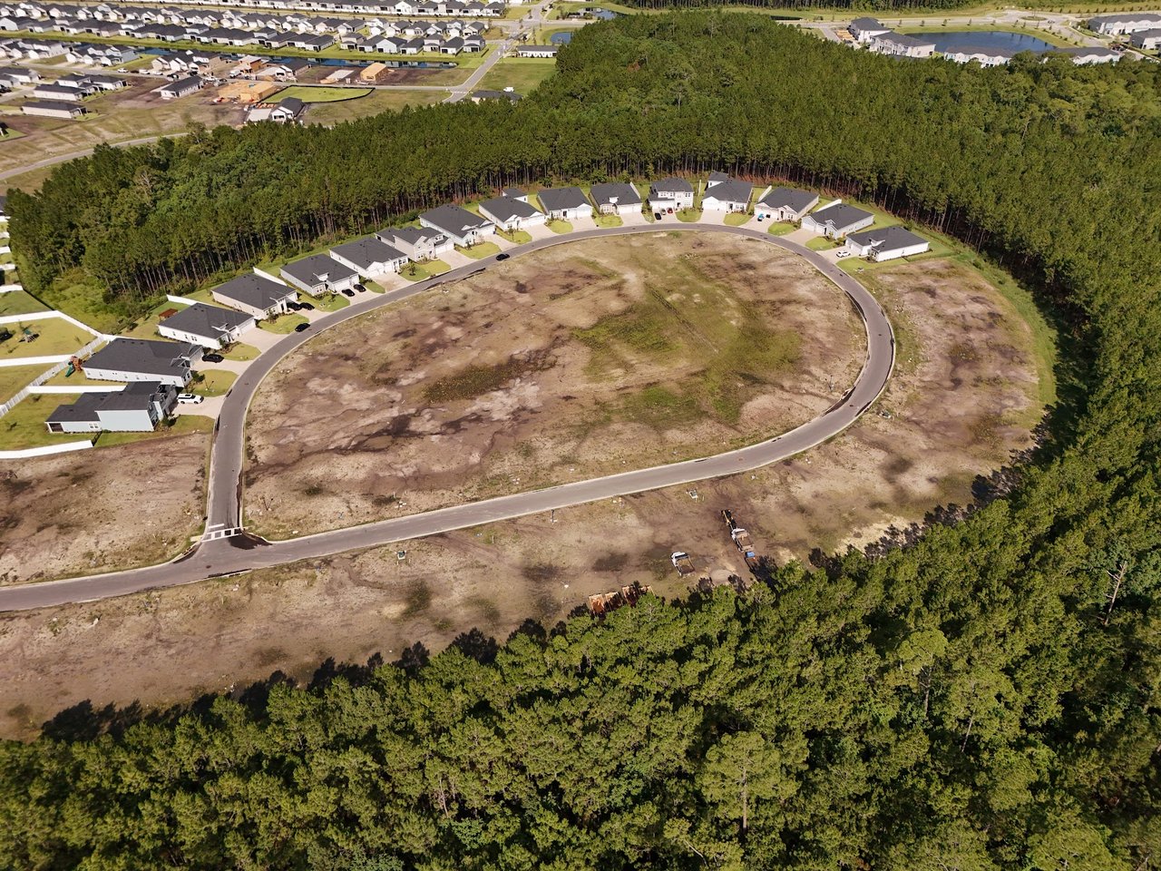 Aerial view of homes along a curved road around a large undeveloped lot, bordered by trees.