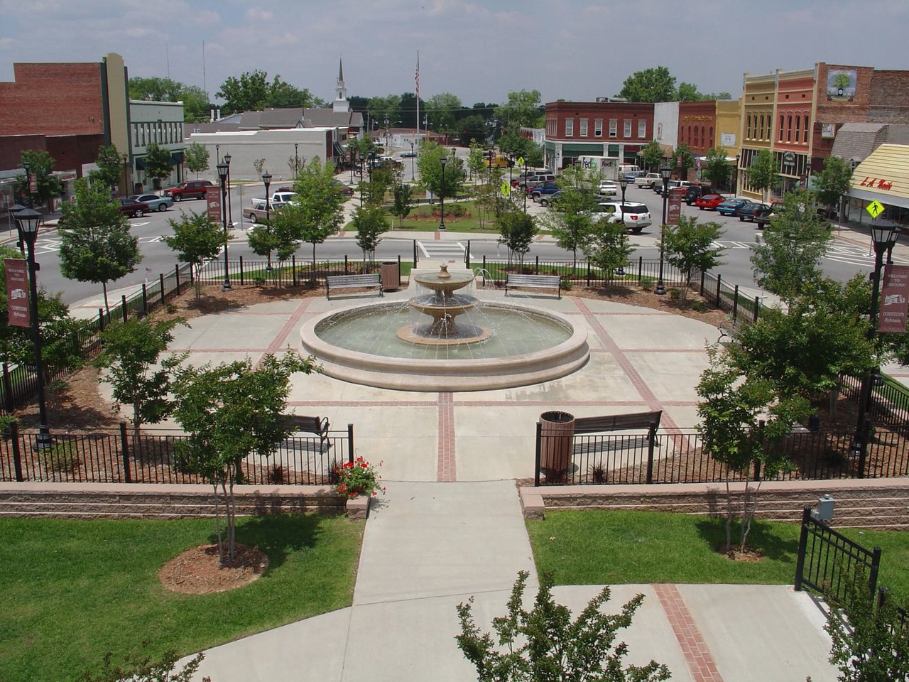 Downtown square with a tiered fountain, benches, and brick storefronts.