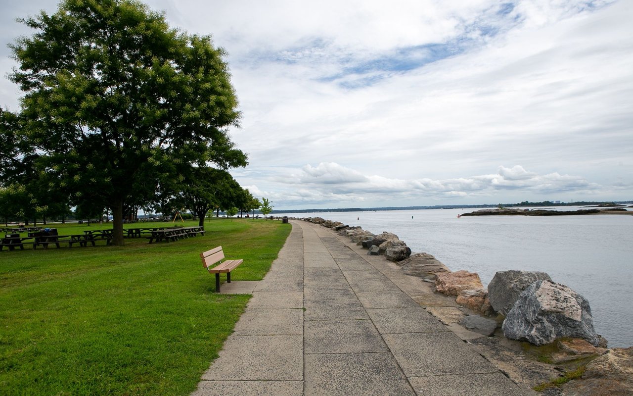Waterfront promenade in New Rochelle with a tree-shaded lawn, bench, and rocky shoreline.