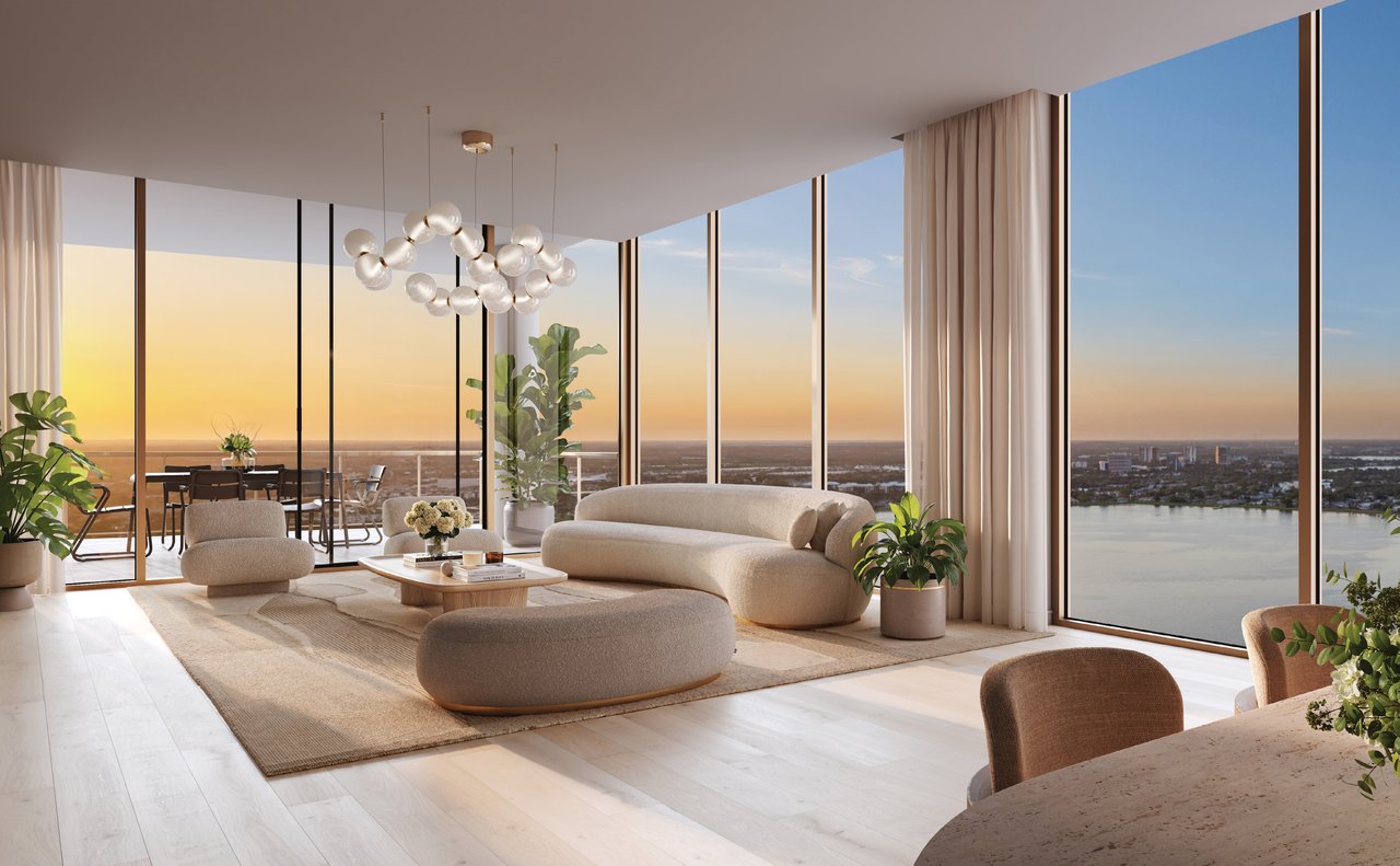 Staged condo living room with curved cream seating and floor-to-ceiling windows overlooking water at sunset.