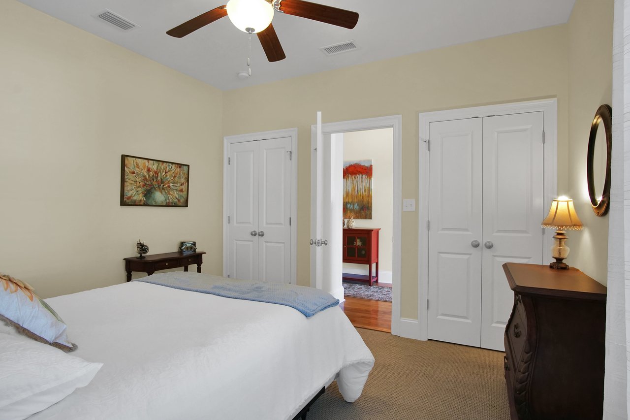 Cozy bedroom with a white bedspread, wooden furniture, and soft lighting. A ceiling fan hangs above, adding a calm, inviting atmosphere.