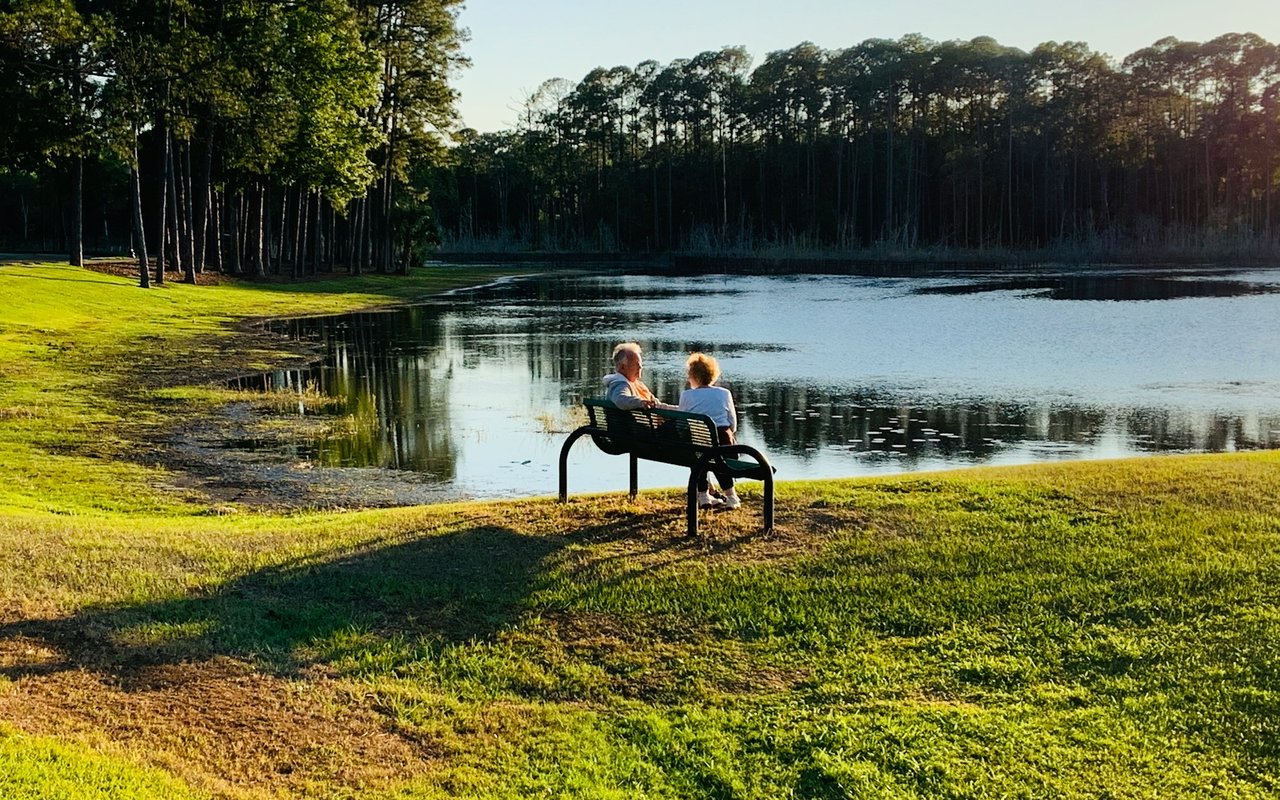 A couple sits on a bench beside a tree-lined lake and grassy shore.