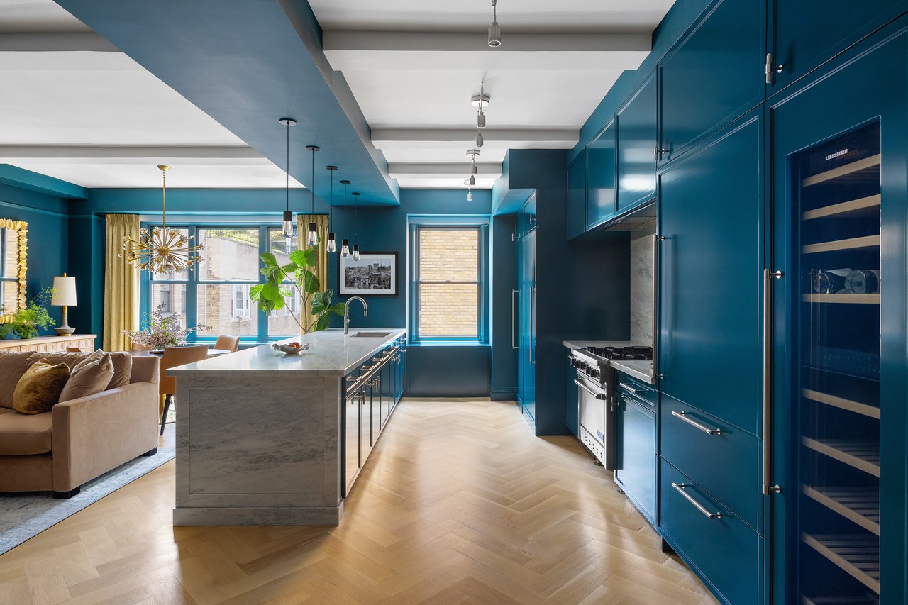 Open-plan kitchen with teal cabinets, a marble island, and a living area beside large windows.