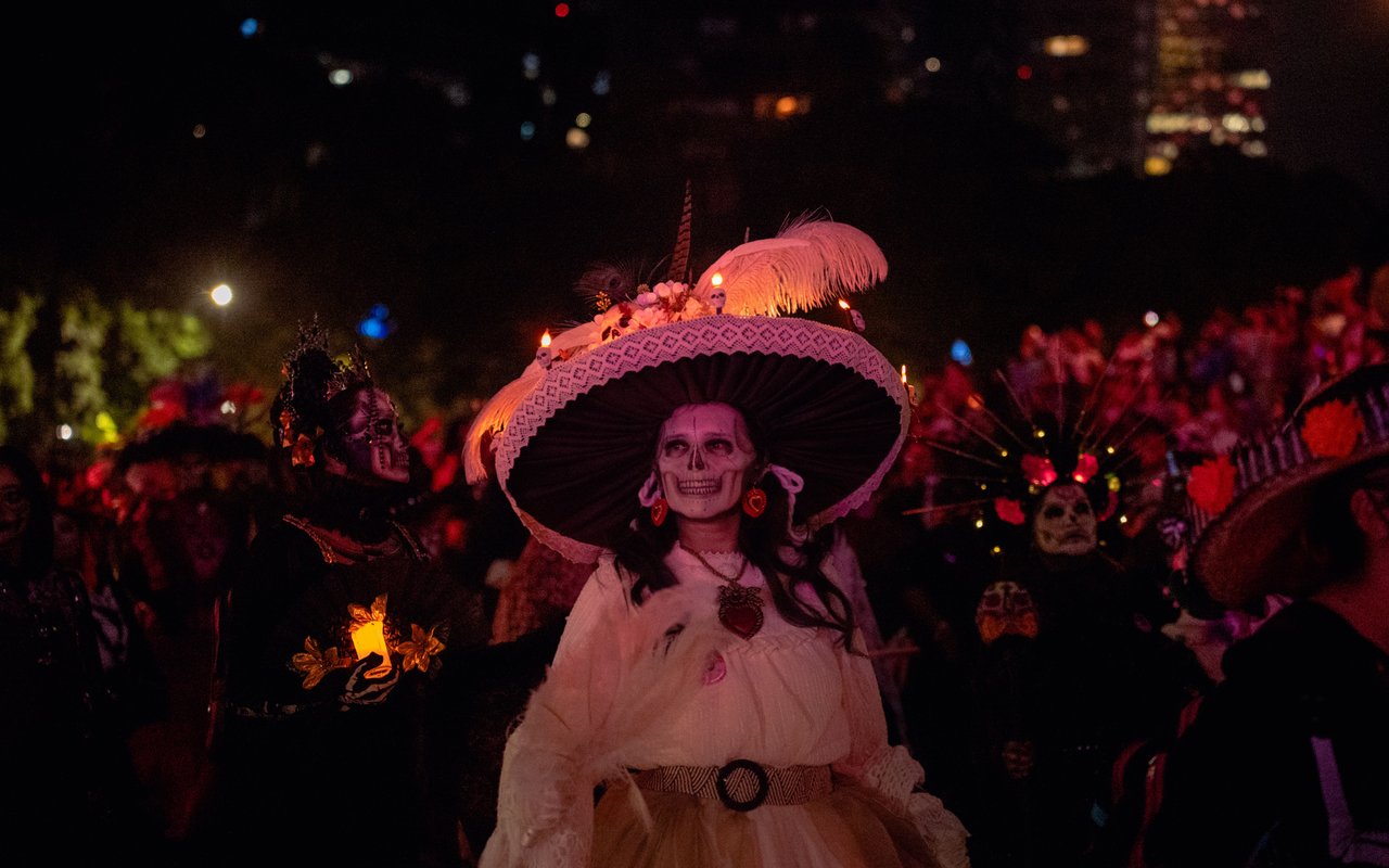 A nighttime photograph of a historic West Village street decorated for Halloween, featuring brick townhouses, pumpkins, costumed pedestrians, police barricades, and the glow of Halloween celebrations near Sixth Avenue.