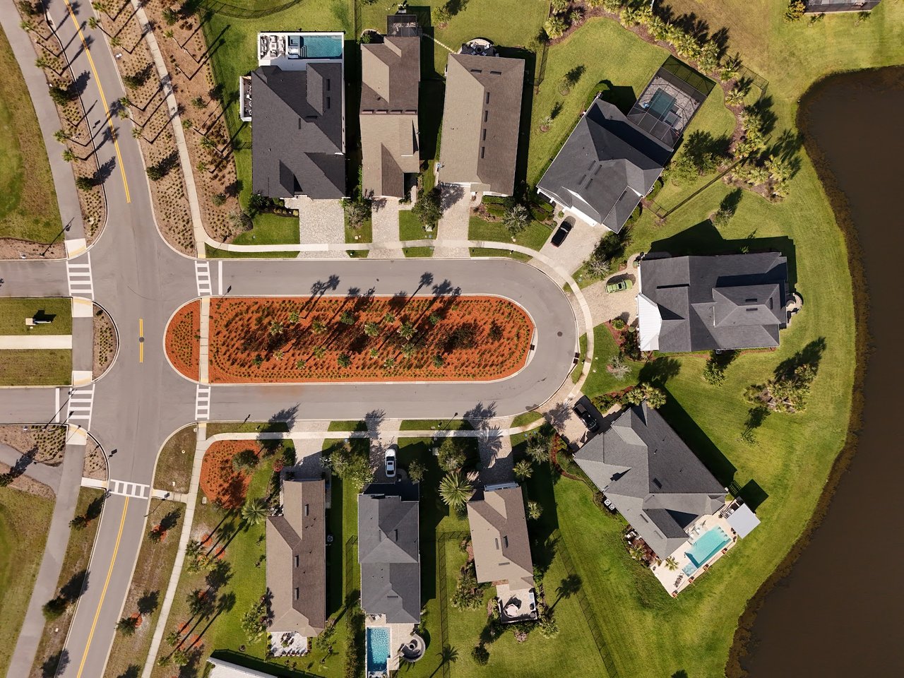 Aerial view of homes around a landscaped cul-de-sac beside a pond, with several private pools.