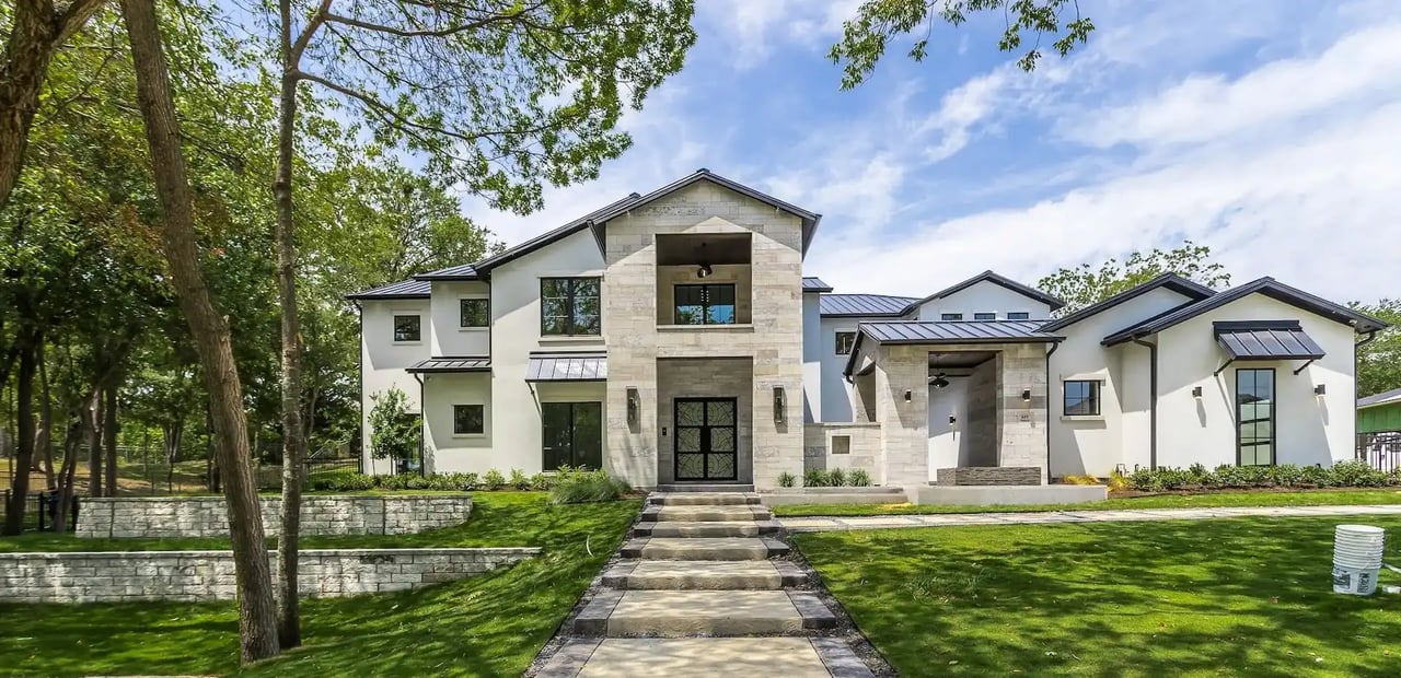 Modern two-story home with a pale stone entrance, dark roof, and broad walkway through a tree-shaded lawn.