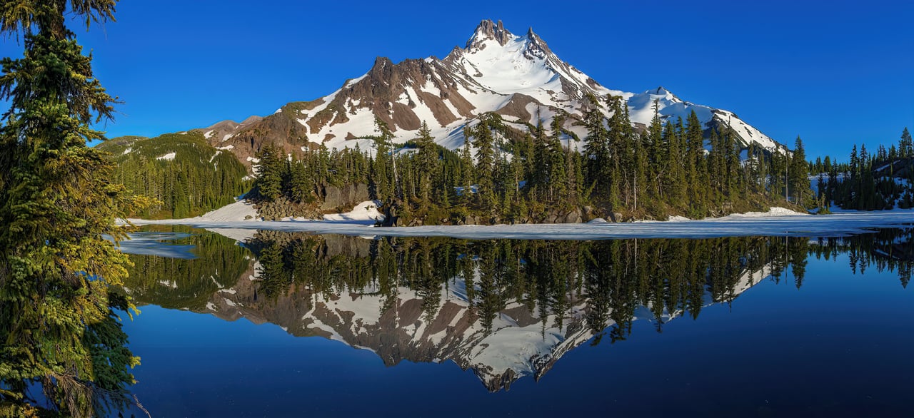 Snow-covered mountain and evergreen forest reflected in a blue alpine lake.