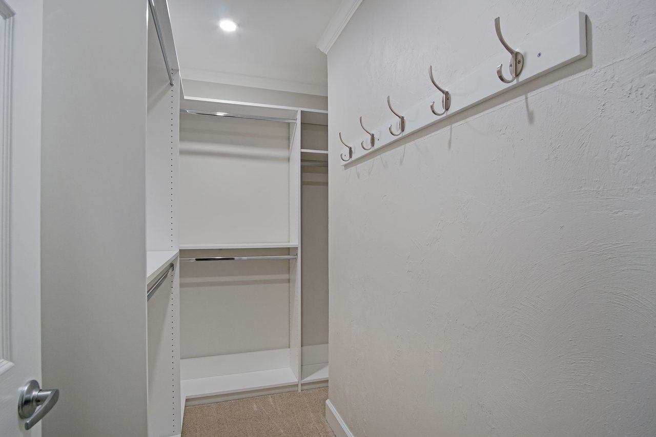 Minimalist walk-in closet with white walls, six hooks on the right, and sleek shelving on the left. Bright overhead lighting creates a clean, organized vibe.