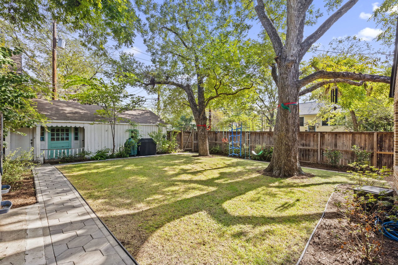 Fenced backyard with mature shade trees, a paved walkway, and a turquoise-door outbuilding.