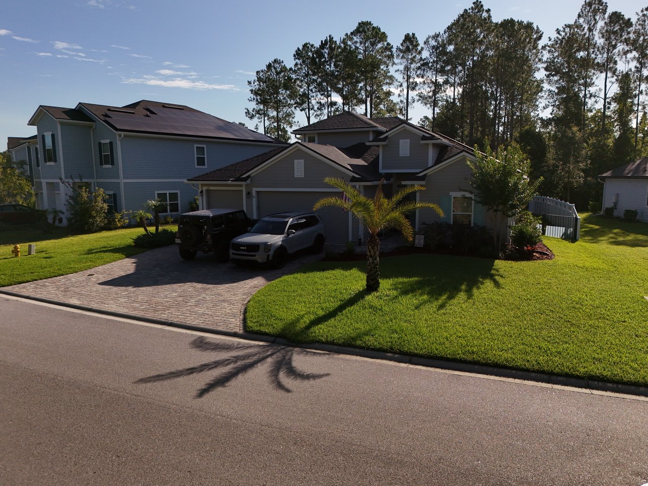 Gray two-story home with rooftop solar panels, a brick driveway, and a palm tree in the front lawn.