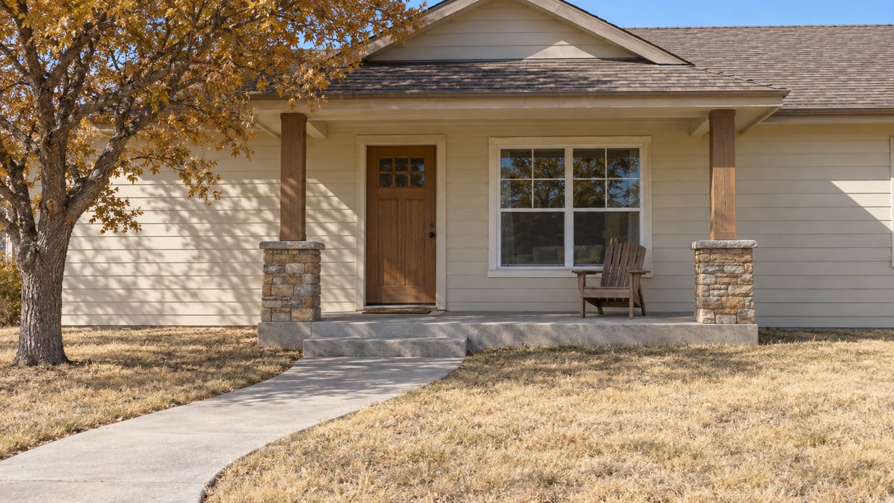 An autumn oak shades the front yard of a cream-sided gabled home with a stone-accented covered porch and a weathered concrete approach.