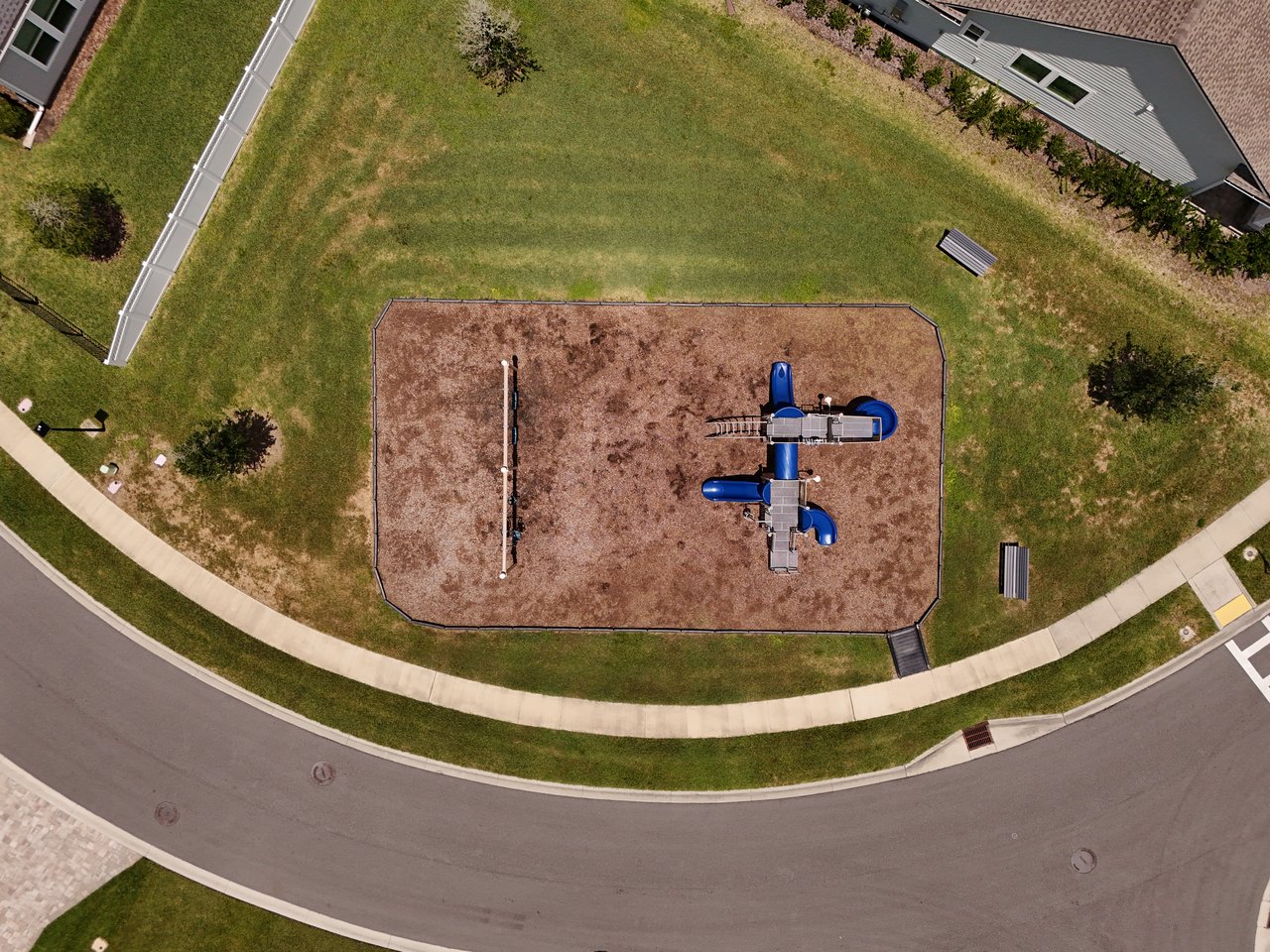 Neighborhood playground with blue play equipment in a fenced mulched area, surrounded by lawns and sidewalks.