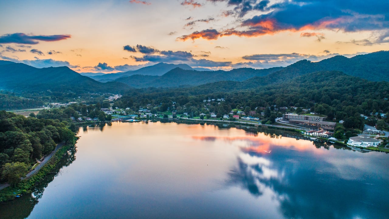 Aerial view of a tranquil lake surrounded by forested hills and a small village at sunset, with a reflection on the platform of the water's surface that real estate agents find enchanting.