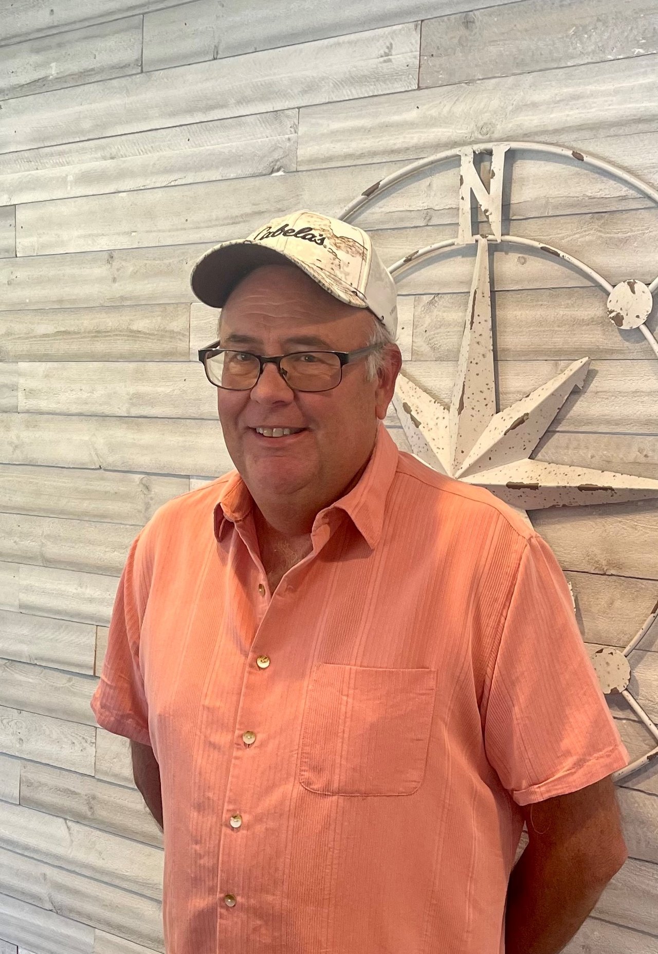Steven Church wearing glasses, a white cap, and a coral shirt in front of a nautical compass.