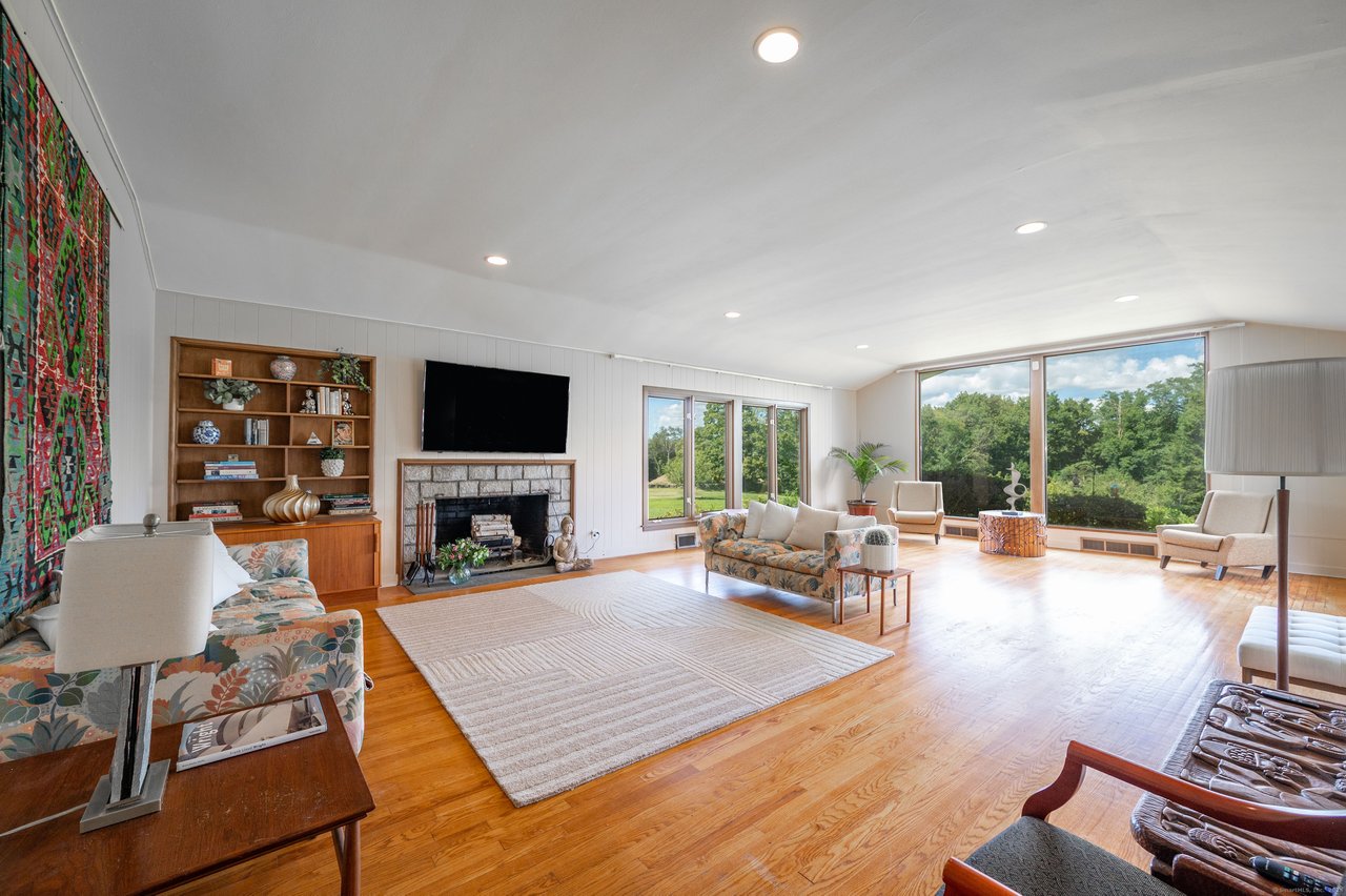 Living room with a stone fireplace, built-in shelves, hardwood floors, and large windows overlooking greenery.
