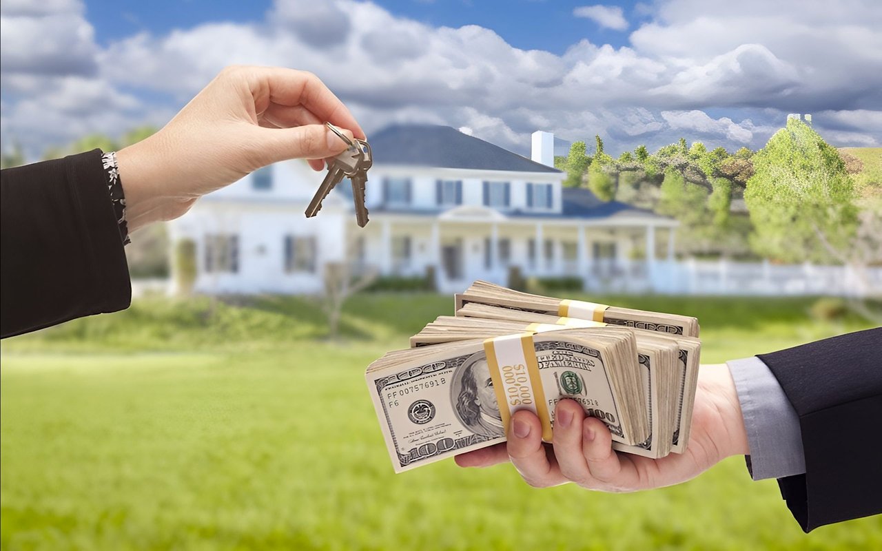 Hands exchange house keys and cash in front of a large white house.