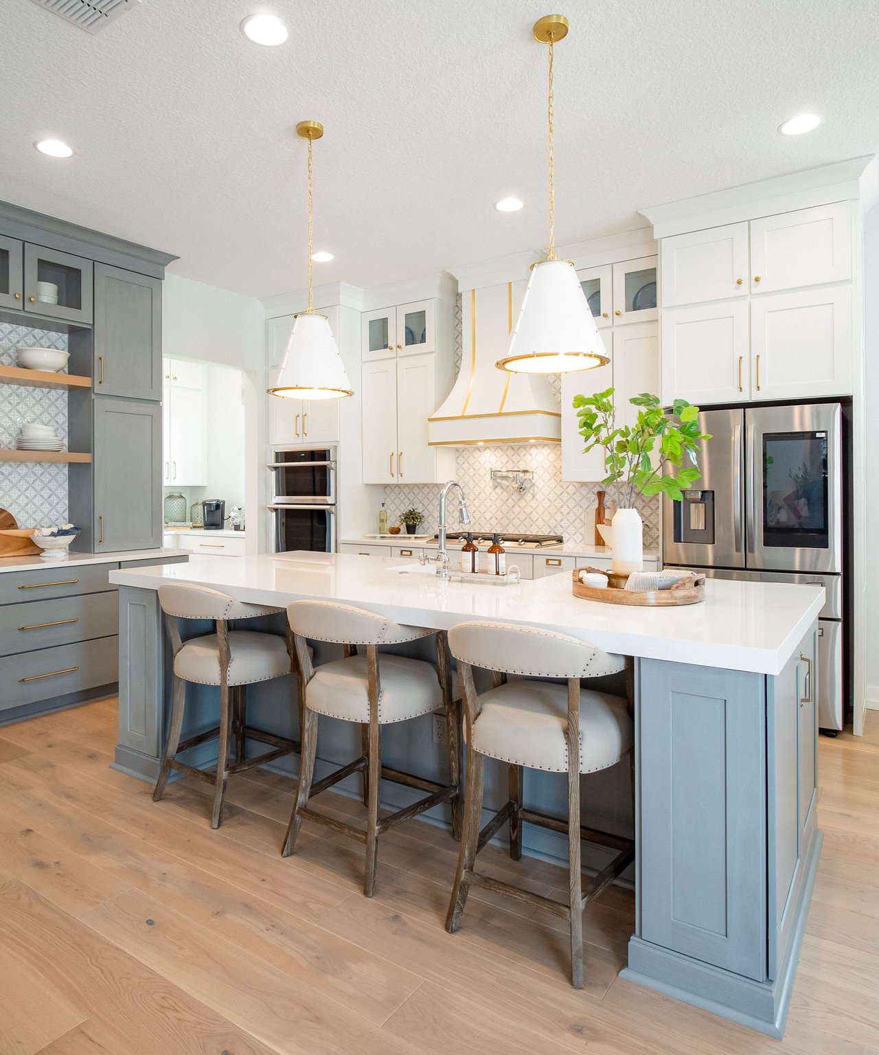 Modern kitchen with a large white island, three bar stools, gray-blue cabinets, and gold pendant lights.