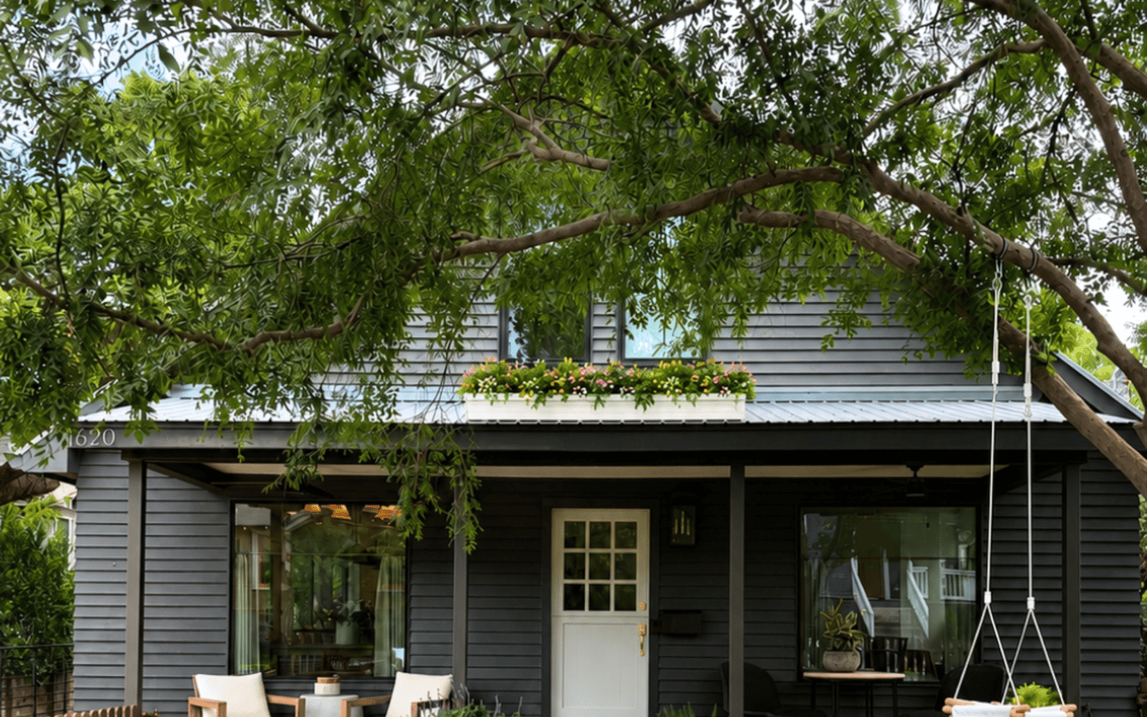 Gray house with a covered porch, flower box, and hanging swing beneath a leafy tree.