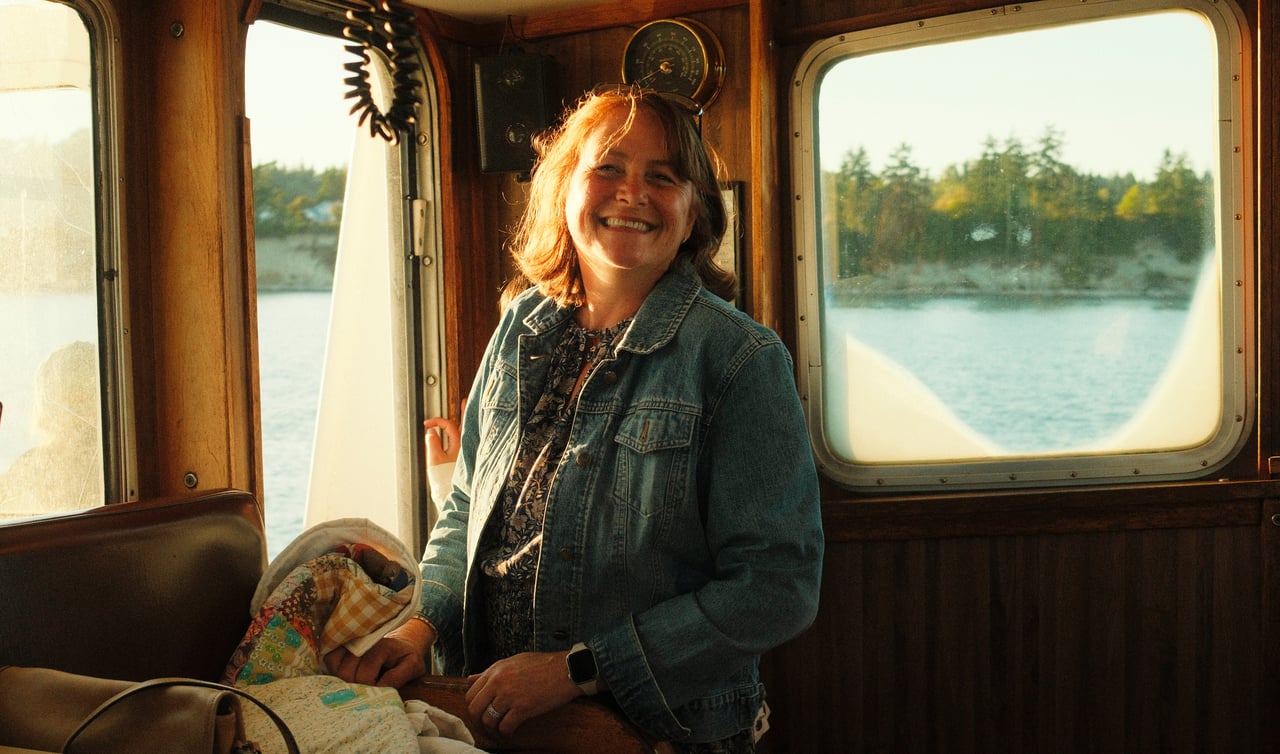 A smiling woman in a denim jacket aboard a wooden boat, with water visible through the windows.