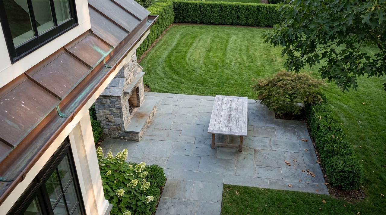Bluestone terrace with a stone fireplace and teak table beside a striped lawn, clipped hedge, and mature trees.