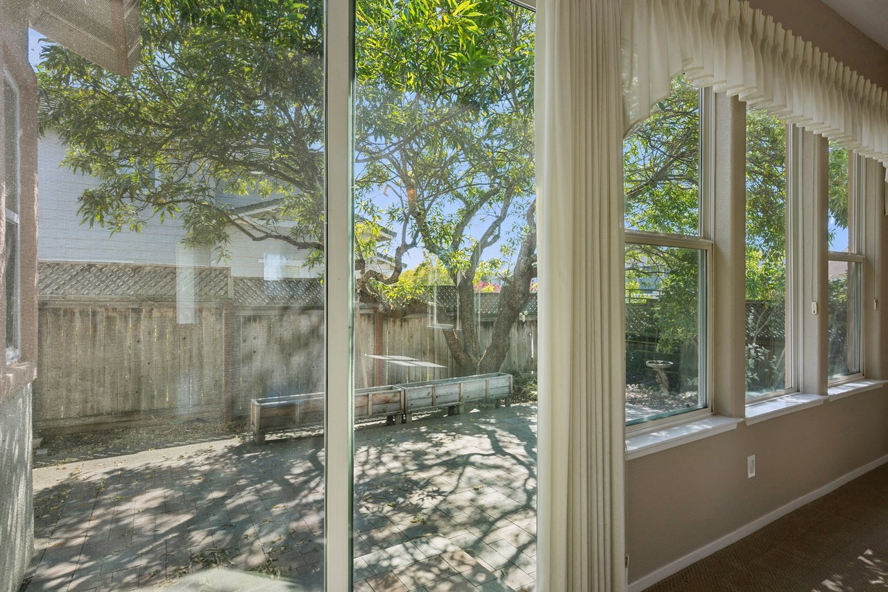 Interior room with tall windows and cream curtains overlooking a tree-shaded backyard patio.