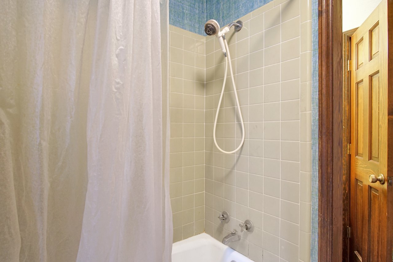 A small bathroom shower with beige tiles, a white showerhead, and a white curtain partially open. A wooden door is slightly ajar, revealing warm lighting.